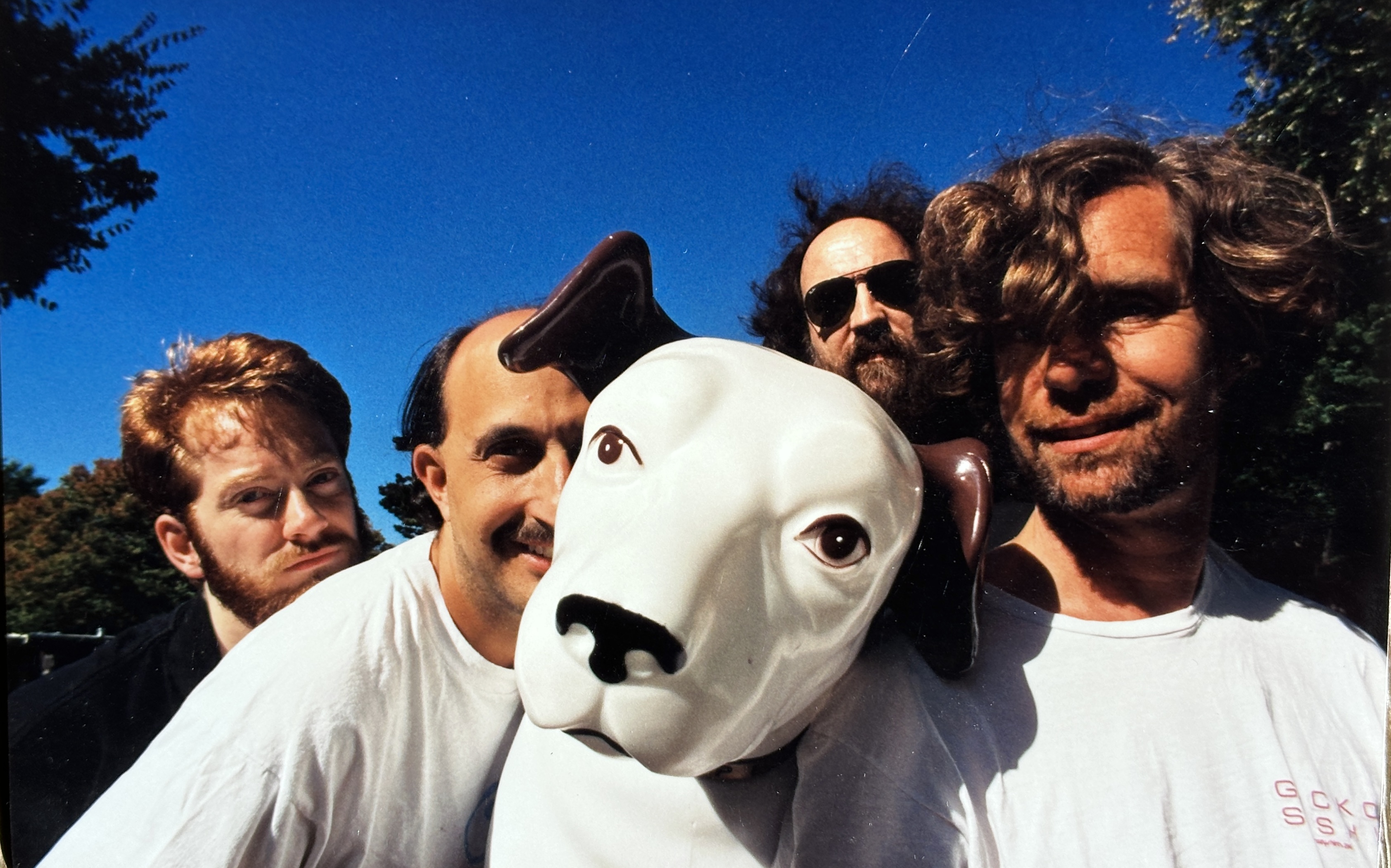 Qwire band photo featuring members gathered around a white dog sculpture under a vivid blue sky.